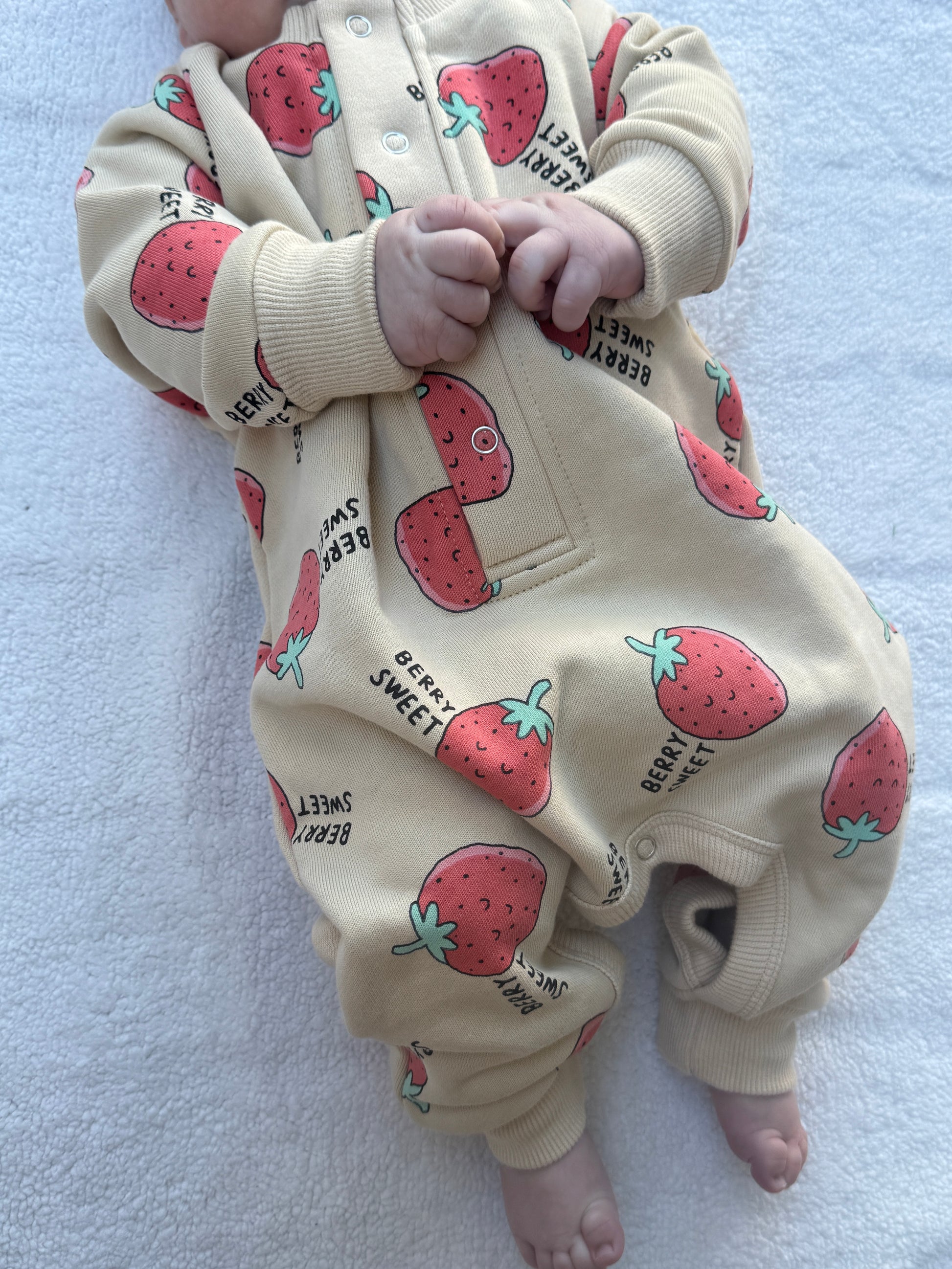 A baby wearing a cream-colored one-piece jumpsuit with a strawberry print, lying on a white textured blanket.