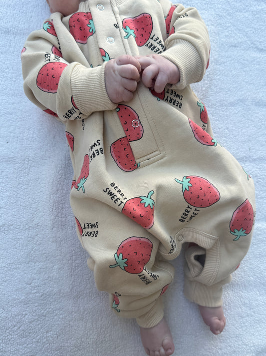 A baby wearing a cream-colored one-piece jumpsuit with a strawberry print, lying on a white textured blanket.